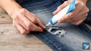 A person using Speed-Sew fabric glue to apply a decorative patch to a pair of denim jeans on a wooden craft table.